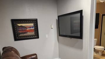 A living room with a brown leather recliner, a framed picture on the wall, and a flat-screen TV mounted above a toilet.