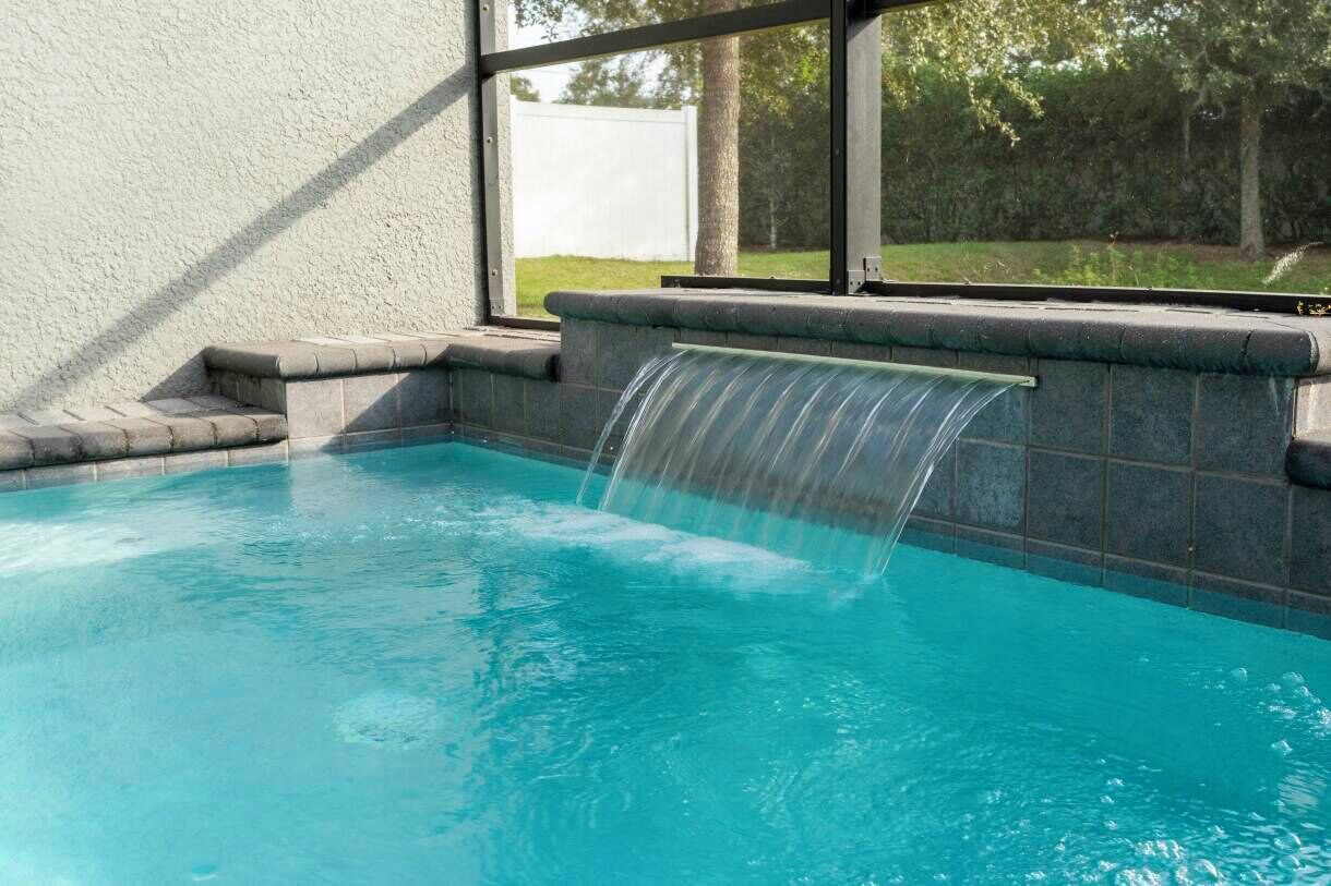 A swimming pool with a waterfall feature, surrounded by a stone wall and a window overlooking a grassy area.
