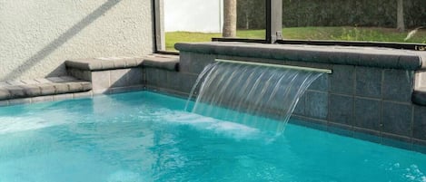 Piscina con cascada, rodeada por un muro de piedra y una ventana con vistas a un espacio cubierto de césped.