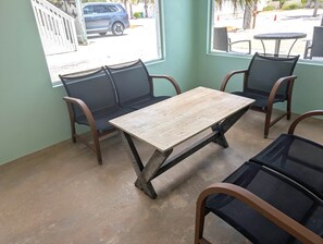 Une pièce avec une table en bois et quatre chaises, une fenêtre donnant sur une voiture garée et une plage, et un sol en béton.