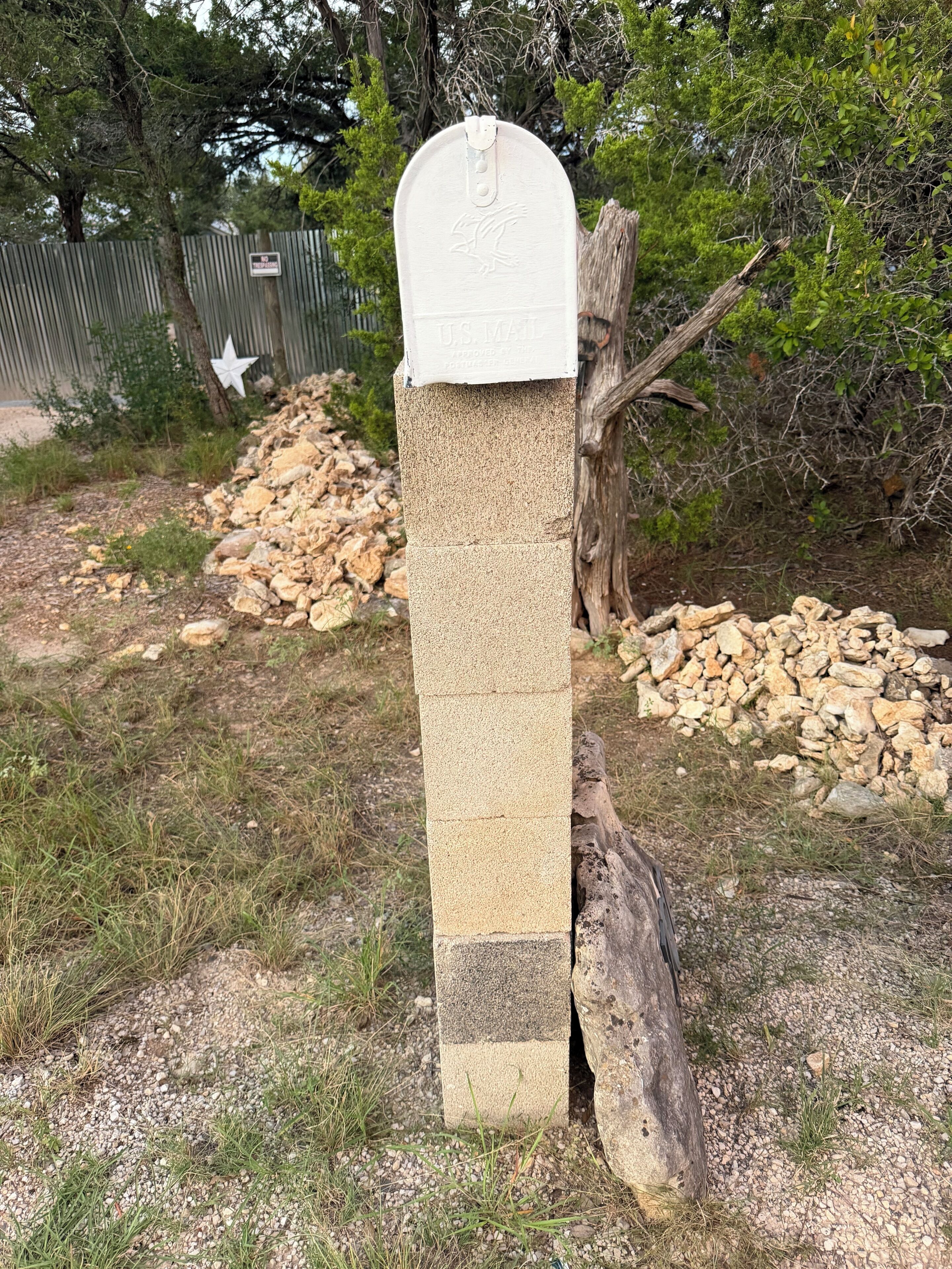 A mailbox with a white post and a U.S. Mail label, surrounded by gravel and vegetation.