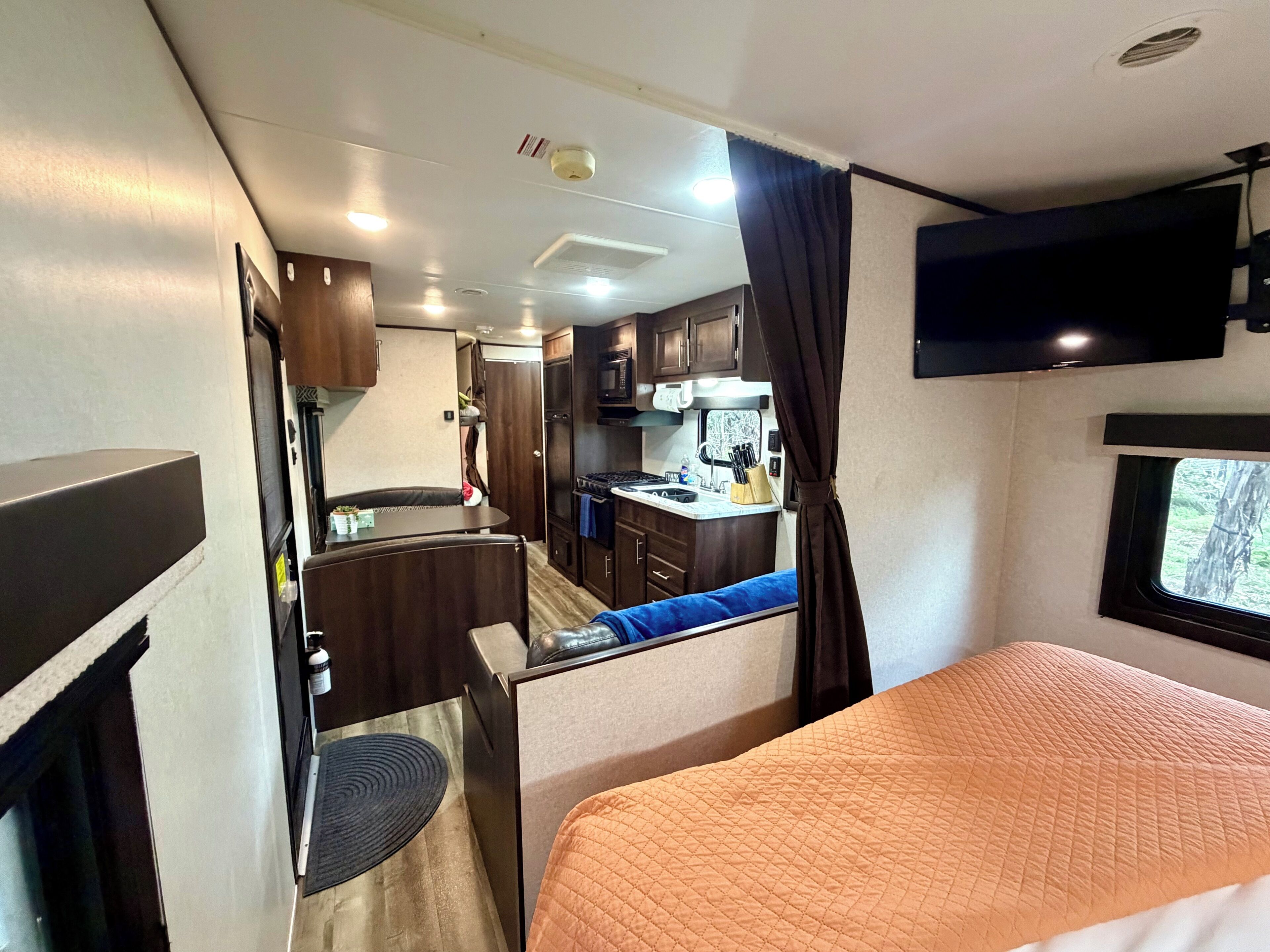A compact RV interior with a kitchenette, a bed, and a TV mounted on the wall.
