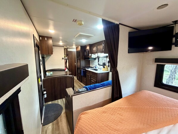 A compact RV interior with a kitchenette, a bed, and a TV mounted on the wall.