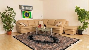A living room with a beige sectional sofa, a glass coffee table, and a patterned rug. There are two potted plants and a framed picture on the wall. - Ein Zimmer in 2 Wohnung Zimmer (Vienna)