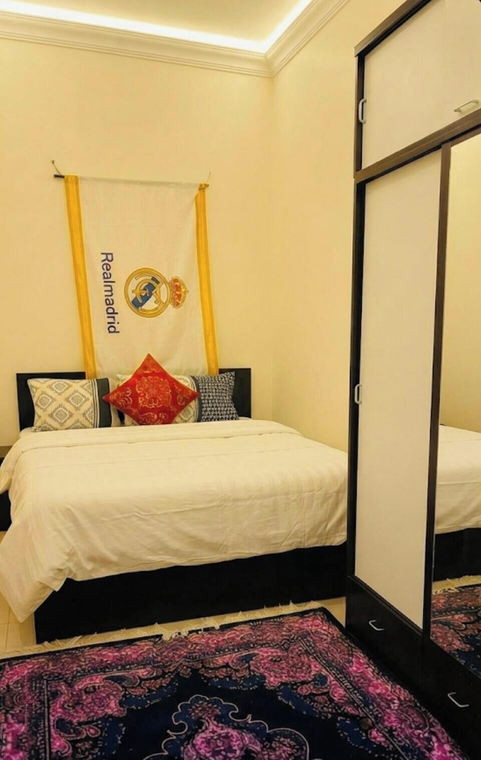 A hotel room with a bed, a mirror, a patterned rug, and a wall hanging with a logo.
