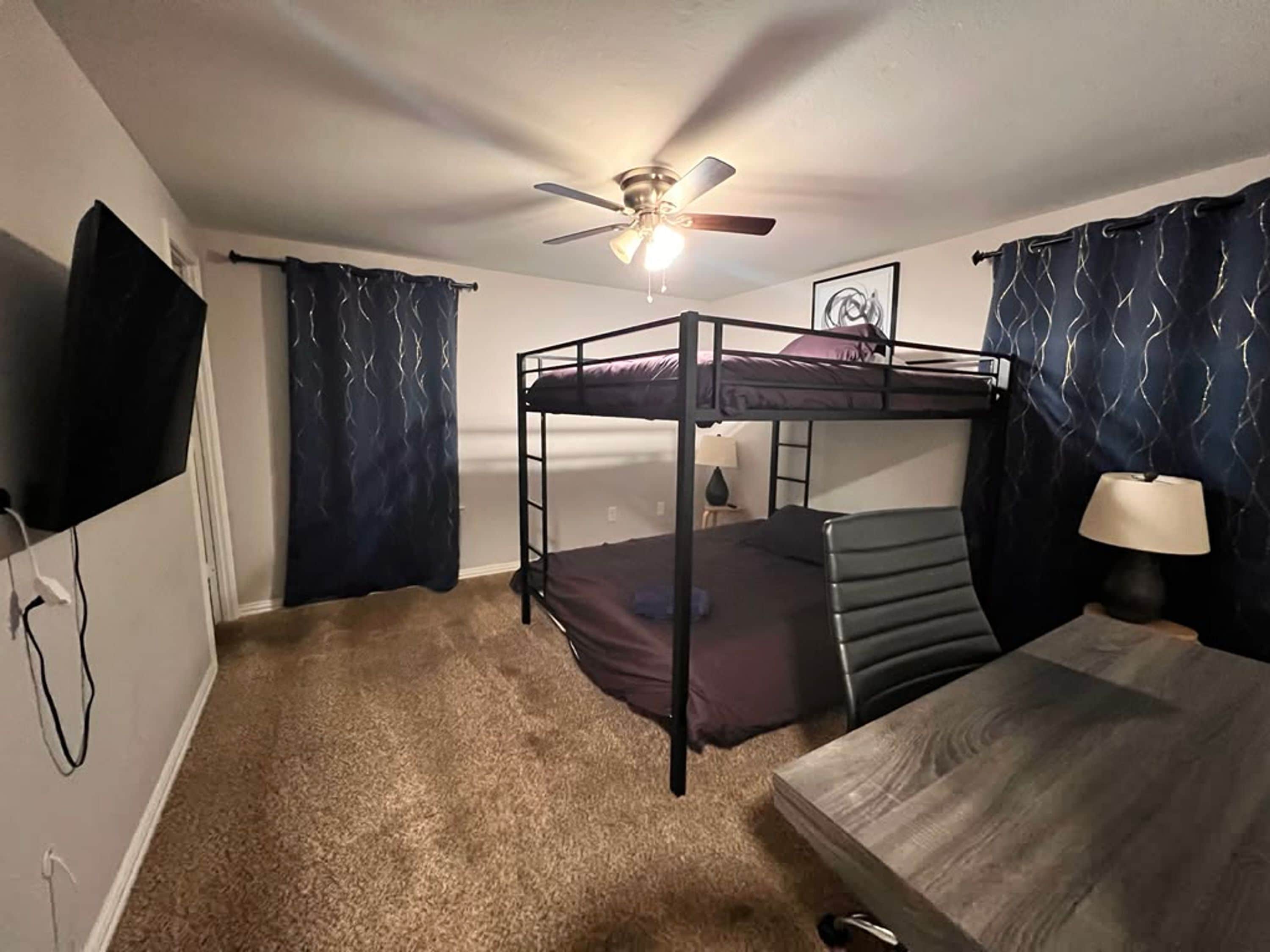 A bunk bed room with a ceiling fan, a TV mounted on the wall, and a bedside table with a lamp.