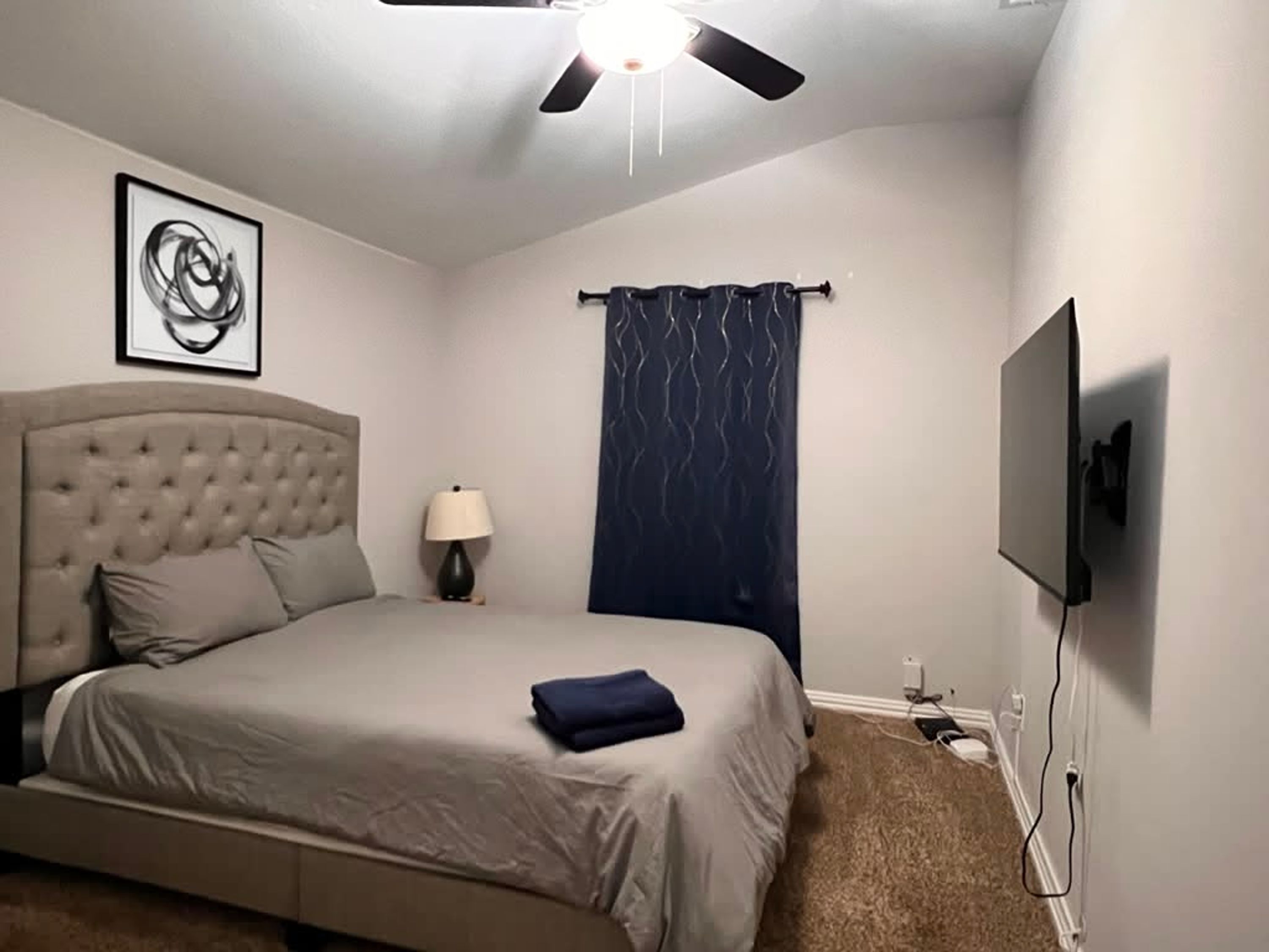 A bedroom with a bed, a ceiling fan, a wall-mounted TV, and a framed picture.