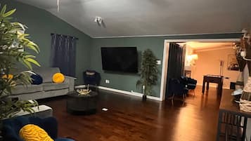 A living room with a sofa, armchair, and coffee table. There is a television mounted on the wall and a plant in the corner.