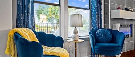 A modern living room with a blue sofa, a yellow throw blanket, and a blue armchair. There is a floor lamp and a view of the outdoors through large windows.