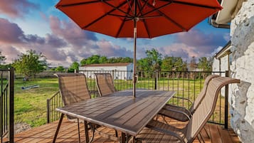 Un patio al aire libre con una mesa y sillas, un paraguas rojo y vistas a una casa y árboles.