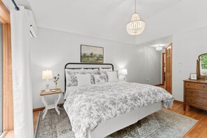 A bedroom with a bed, bedside lamps, a chandelier, a dresser, and a painting on the wall.