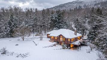A snow-covered house with a chimney emitting smoke, surrounded by trees and a snowy landscape.