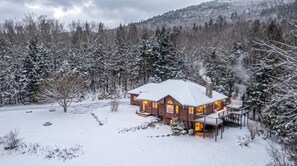 A snow-covered house with a chimney emitting smoke, surrounded by trees and a snowy landscape. - Private 2-Acre Escape | Hot Tub • Fire Pit • Games (Bartlett)