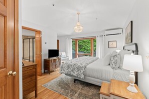Ein Schlafzimmer mit Bett, Nachttisch, Lampe, Fernseher, Kommode und Blick auf Grünfläche.
