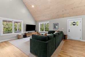 Ein modernes Wohnzimmer mit einem Ecksofa, einem Flachbildfernseher und großen Fenstern mit Blick auf viel Grün.
