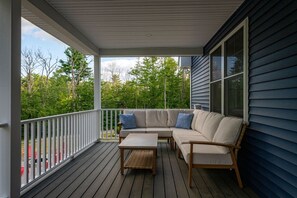 Eine überdachte Veranda mit einer Couch und einem Tisch, mit Blick auf ein bewaldetes Gebiet.