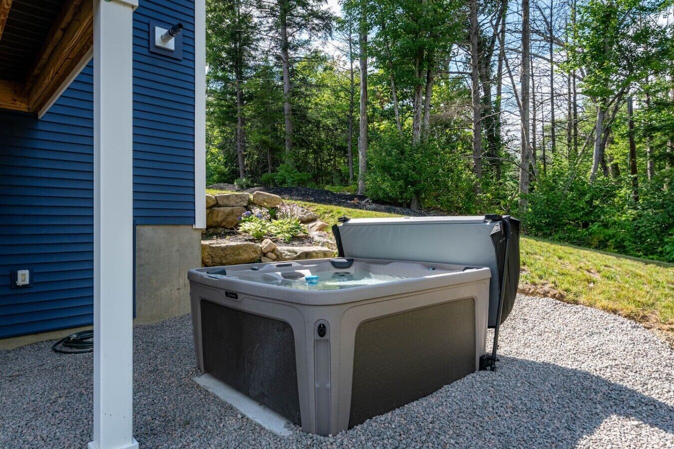 An outdoor hot tub with a stone surround, positioned on a gravel area adjacent to a building with blue siding.