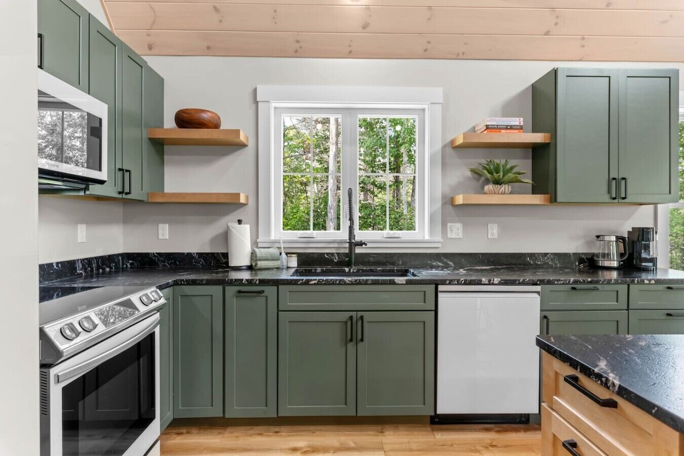A kitchen with green cabinets, a white dishwasher, a black countertop, and a window with a view of trees.