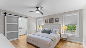 Um quarto com cama, ventilador de teto, janela com persianas e mesa de cabeceira com abajur.