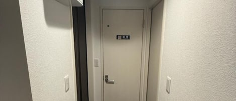 A narrow hallway with a white door at the end, a light fixture on the ceiling, and a small vent on the wall.