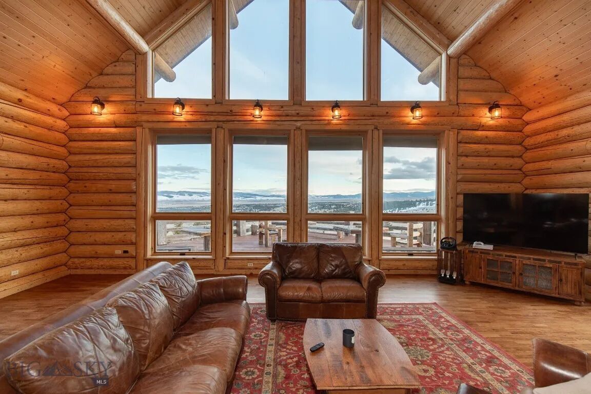 L’intérieur d’une cabane en rondins, avec une grande fenêtre, un canapé en cuir, une table basse en bois et un téléviseur.