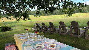 An outdoor dining setup with a table set for a meal, surrounded by chairs, in a grassy field with trees in the background. - Historic 1789 Luxury Farmhouse near Dartmouth College (Hartford)