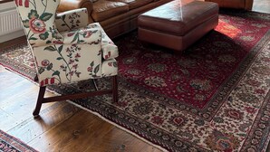 A room with a brown leather sofa, floral armchair, and ottoman, a red patterned rug, wooden beams on the ceiling, and two windows with views of greenery and a building. - Historic 1789 Luxury Farmhouse near Dartmouth College (Hartford)
