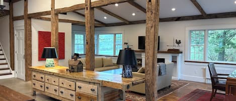 A spacious living area with exposed wooden beams, a central wooden table with drawers, a sofa, and a television mounted on the wall.
