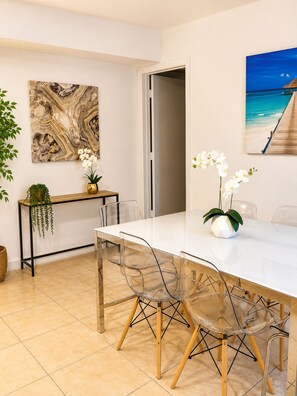 Un coin repas avec une table blanche, des chaises transparentes et un tableau représentant une scène de plage.