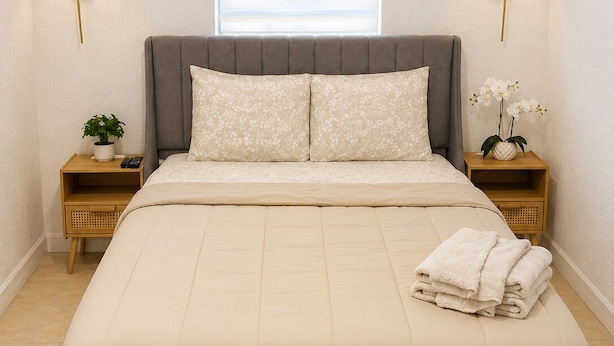 A bed with a beige quilt, two bedside tables with lamps, a window with blinds, and a small potted plant on each table. - Beautiful & Central Cozy Home (Miami)