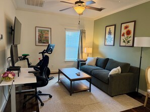 Ein Wohnzimmer mit Schreibtisch, Stuhl, Heimtrainer und einer Couch.
