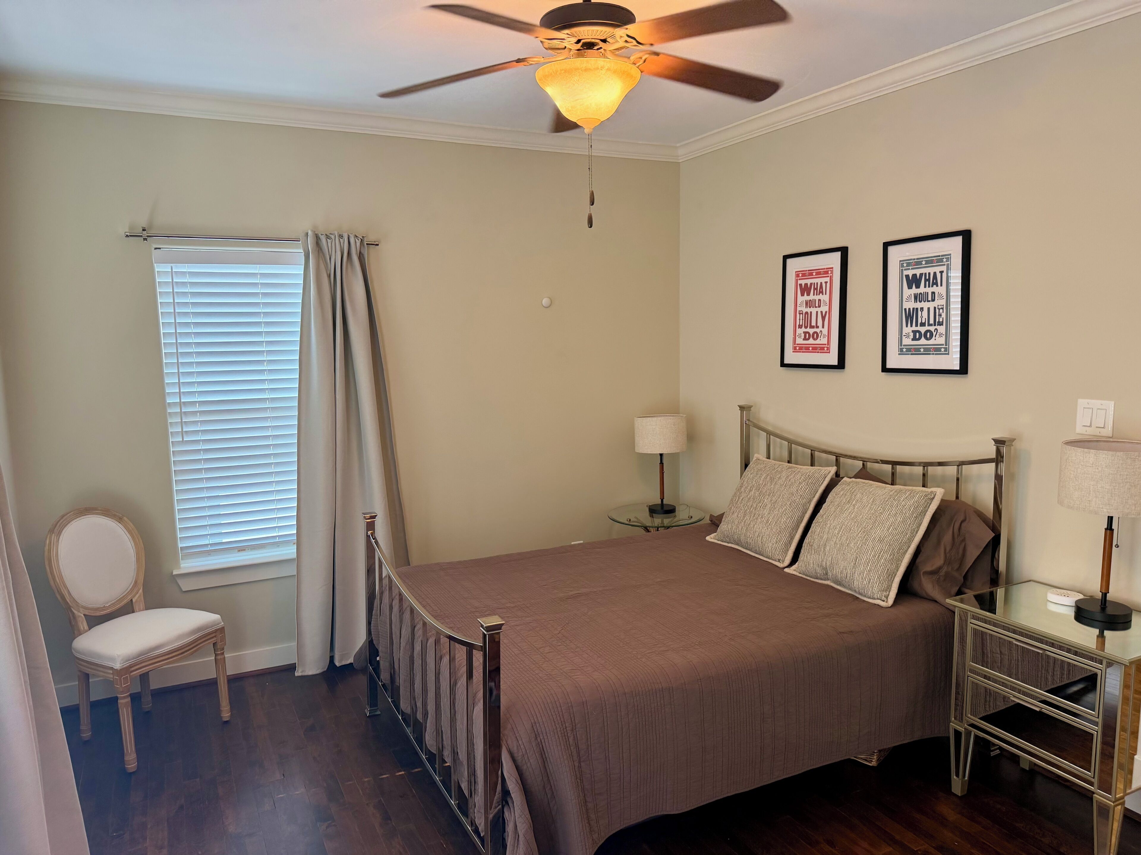 A bedroom with a bed, a chair, a nightstand, a lamp, a ceiling fan, and two framed pictures on the wall.