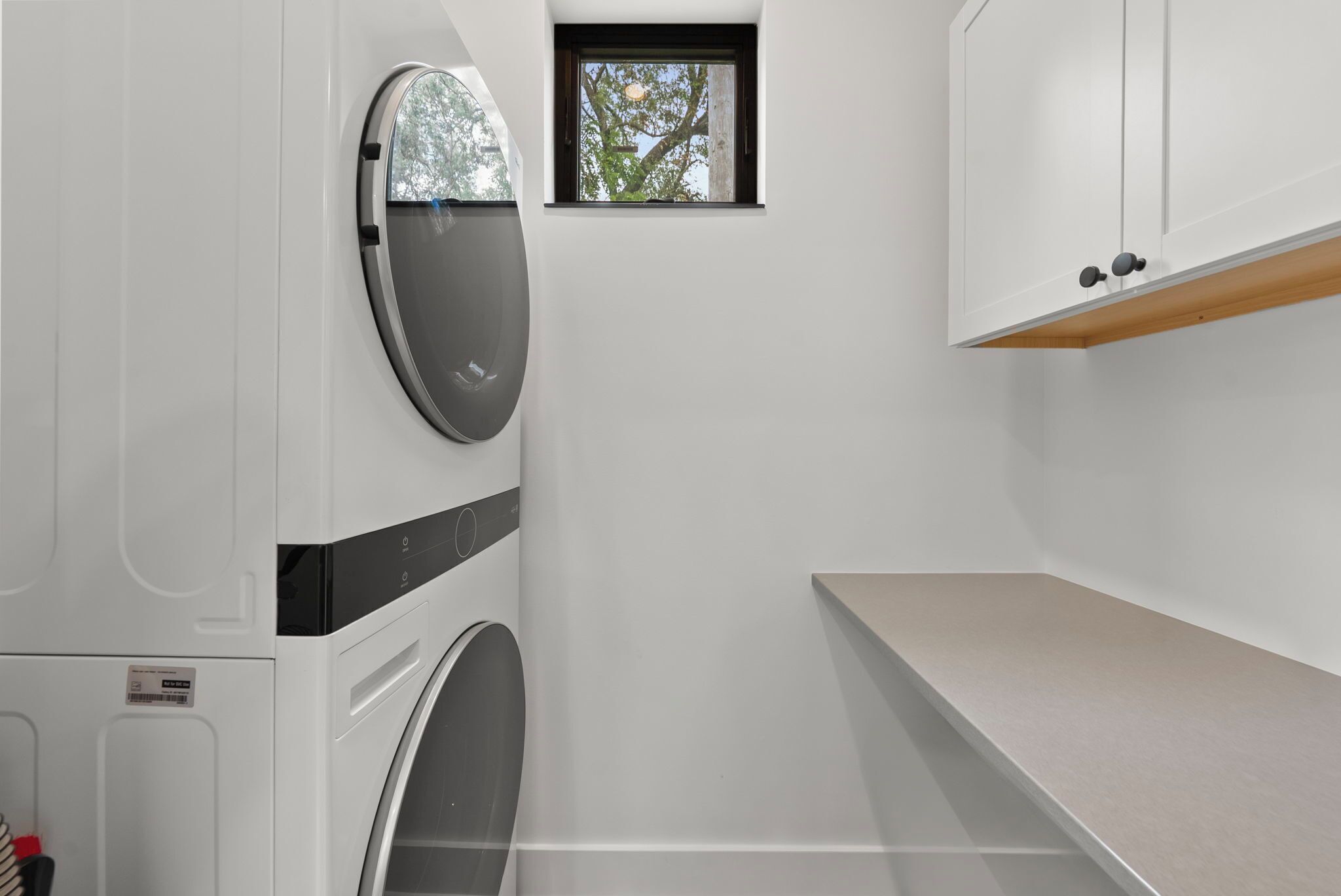 A laundry room with two front-loading washing machines, a small window with a view of trees, and a wooden shelf above a countertop.