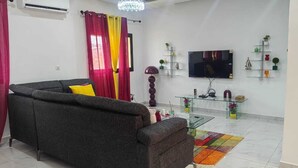 A living room with a sofa, a glass coffee table, a flat-screen TV, and decorative items on shelves. - 2-bedroom Residences meublees in soothing Bouake with AC comfort (Bouake)