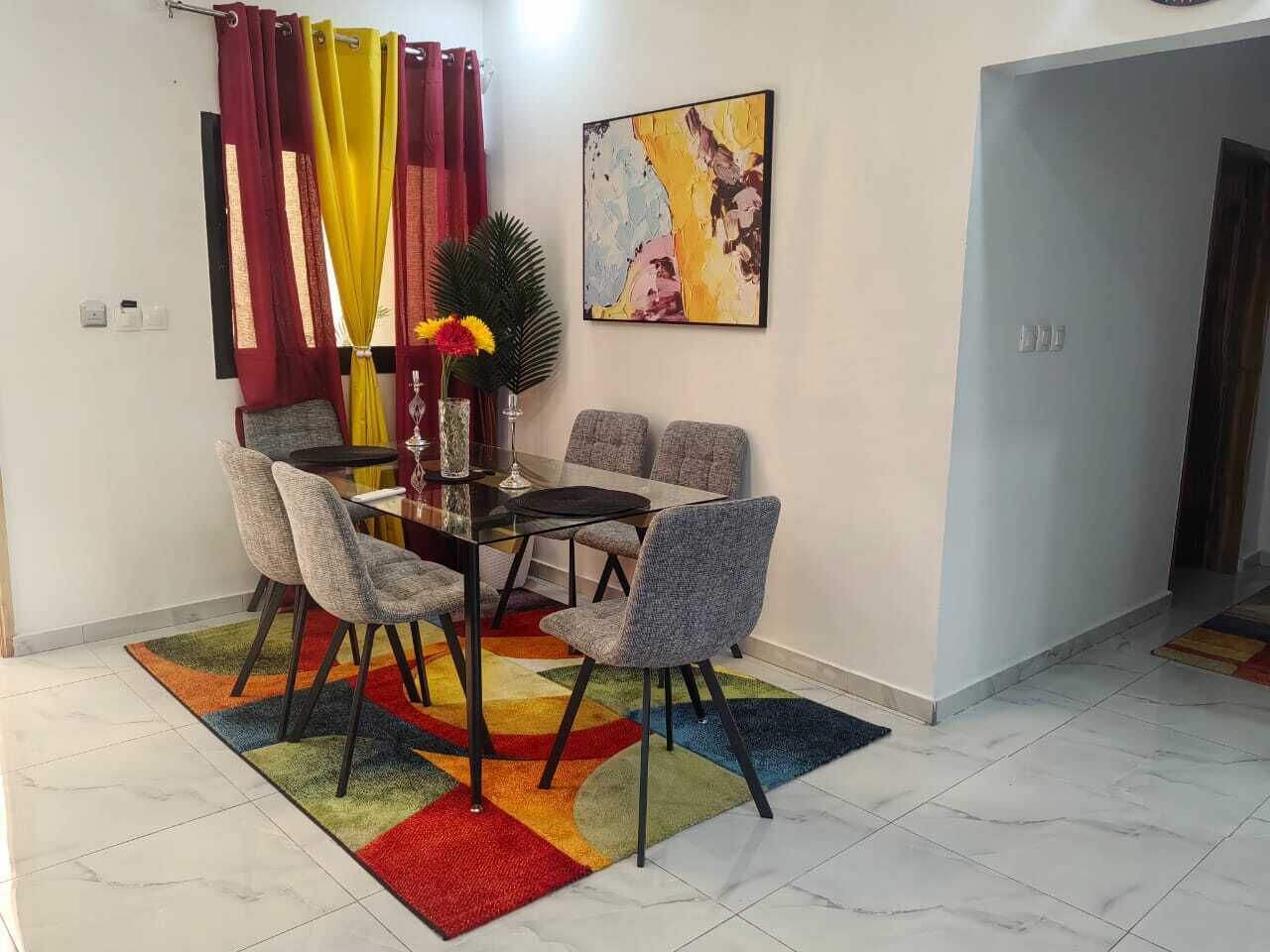 A dining area with a glass table and grey upholstered chairs, a colorful rug, and a painting on the wall.