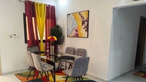A dining area with a glass table and grey upholstered chairs, a colorful rug, and a painting on the wall. - 2-bedroom Residences meublees in soothing Bouake with AC comfort (Bouake)