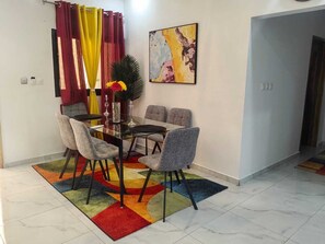 A dining area with a glass table and grey upholstered chairs, a colorful rug, and a painting on the wall. - 2-bedroom Residences meublees in soothing Bouake with AC comfort (Bouake)