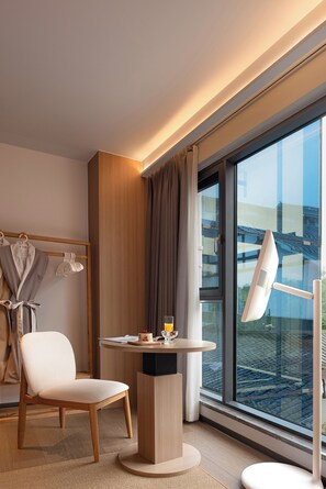 A hotel room with a large window, a round table, a chair, a robe rack, and a desk with a lamp. - Zhangjiajie Ruyuan Hotel  (Zhangjiajie)