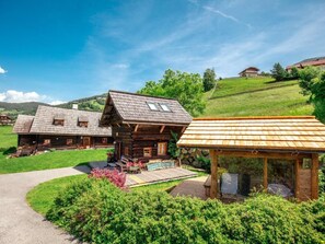 En træhytte med veranda og overdækket siddeområde, omgivet af frodig vegetation og en klar himmel.
