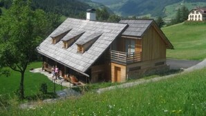 A wooden cabin with a sloped roof, a balcony, and a chimney, surrounded by a lush green landscape and mountains in the background. - Napoleonvilla - Ferienhaus für kreative Auszeiten by Interhome (Rennweg am Katschberg)