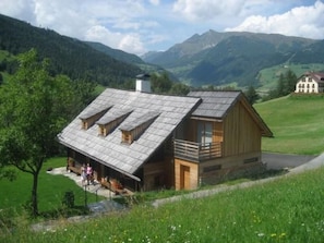 A wooden cabin with a sloped roof, a balcony, and a chimney, surrounded by a lush green landscape and mountains in the background. - Napoleonvilla - Ferienhaus für kreative Auszeiten by Interhome (Rennweg am Katschberg)
