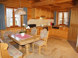 Ein Interieur einer Holzhütte mit Esstisch, Stühlen, einer Bank, einer Küchenzeile und einem Fenster mit Vorhängen.