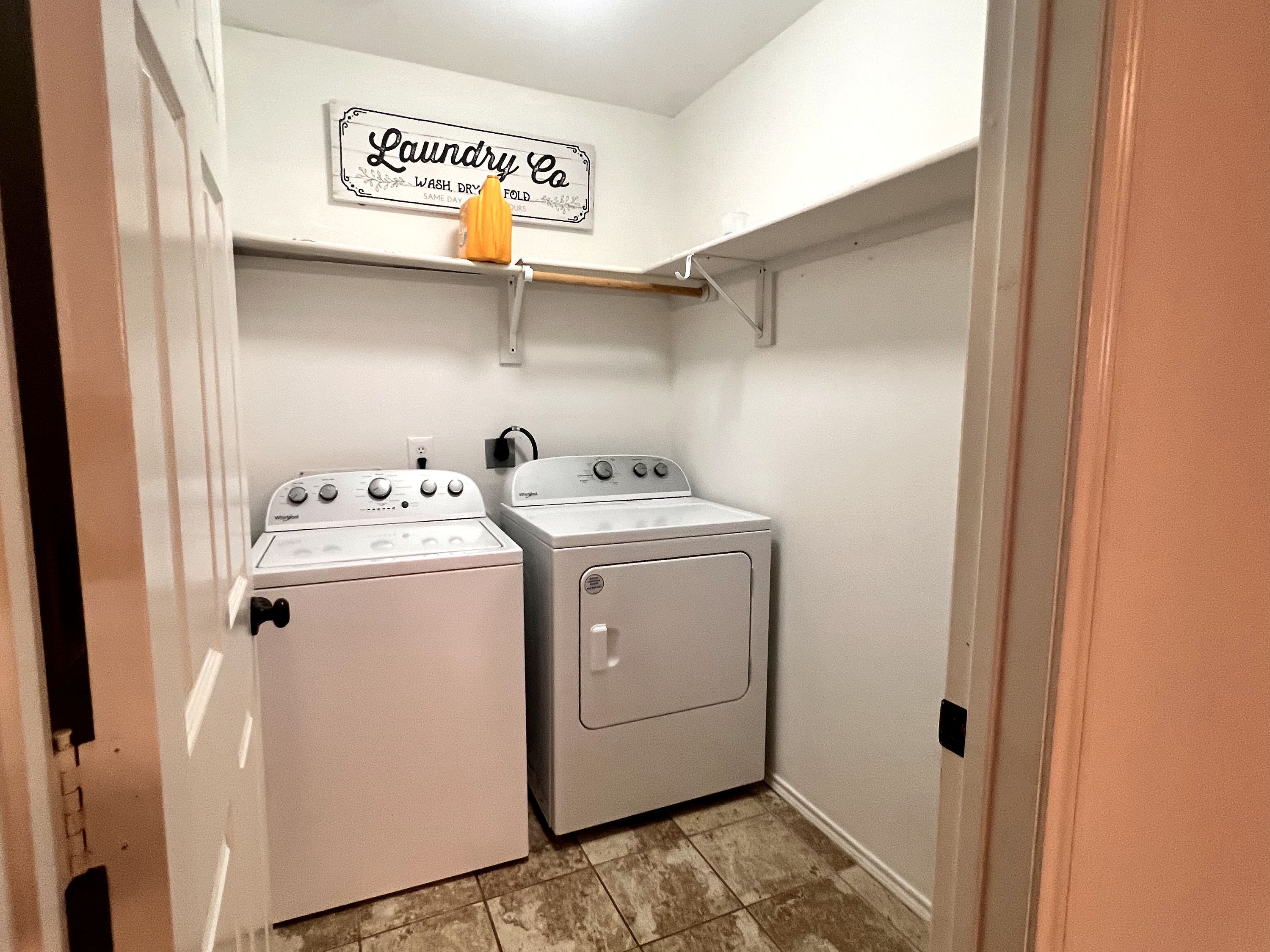 A laundry room with two washers and a dryer, a sign above the dryers, and a small shelf with a cup.