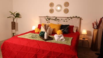 A neatly made bed with a red quilt, a white vase with dried plants, and a small wooden tray.