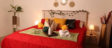 Un lit bien fait, recouvert d’une courtepointe rouge, un vase blanc contenant des plantes séchées et un petit plateau en bois.