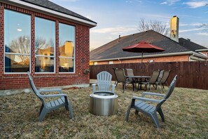 Un patio trasero con un brasero, dos sillas Adirondack, una mesa y un paraguas rojo.