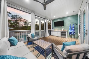 A covered outdoor deck with a sofa, a table, and a kitchen area with a TV. - The Emerald at 30A: Heated Pool, Golf Cart and Bikes INCLUDED (Santa Rosa Beach)