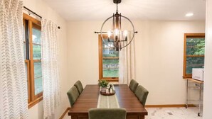 A dining room with a marble floor, a wooden table, green upholstered chairs, a chandelier, and a view of greenery through the windows. - Midtown Omaha Near Everything! No Cleaning Fee! (Omaha)
