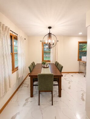 A dining room with a marble floor, a wooden table, green upholstered chairs, a chandelier, and a view of greenery through the windows. - Midtown Omaha Near Everything! No Cleaning Fee! (Omaha)