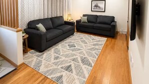 A living room with two black sofas, a patterned rug, wooden flooring, a ceiling fan, and a wall-mounted TV. - Midtown Omaha Near Everything! No Cleaning Fee! (Omaha)